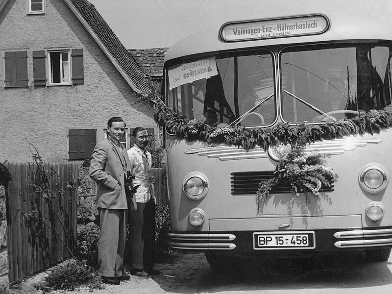 Zwei Männer stehen vor einem mit Girlanden geschmückten Bus, der vor einem Haus geparkt ist.