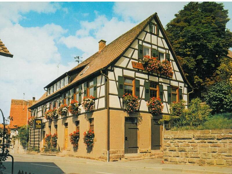 Ein traditionelles Fachwerkhaus mit zahlreichen blühenden Blumenkästen an den Fenstern steht neben einer gepflasterten Straße unter einem blauen Himmel.