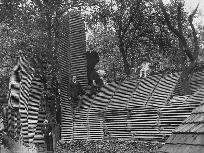 Eine Gruppe von Menschen sitzt auf einem kunstvoll gestapelten Holzstapel, der an eine Skulptur erinnert, umgeben von Bäumen.