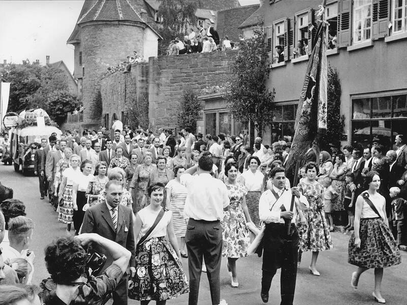 Eine große Menschenmenge in traditioneller Kleidung marschiert in einem Festumzug durch eine Stadt, während Zuschauer von der Seite zusehen.