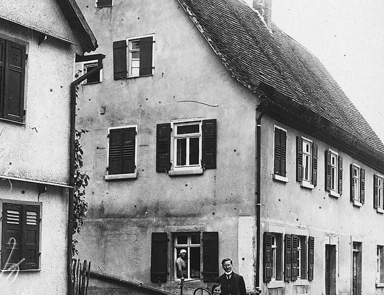 Ein Schwarz-Weiß-Foto zeigt ein kleines Auto und drei Personen vor einem traditionellen mehrstöckigen Haus, wobei eine Person aus einem Fenster schaut und zwei Personen vor dem Auto stehen.