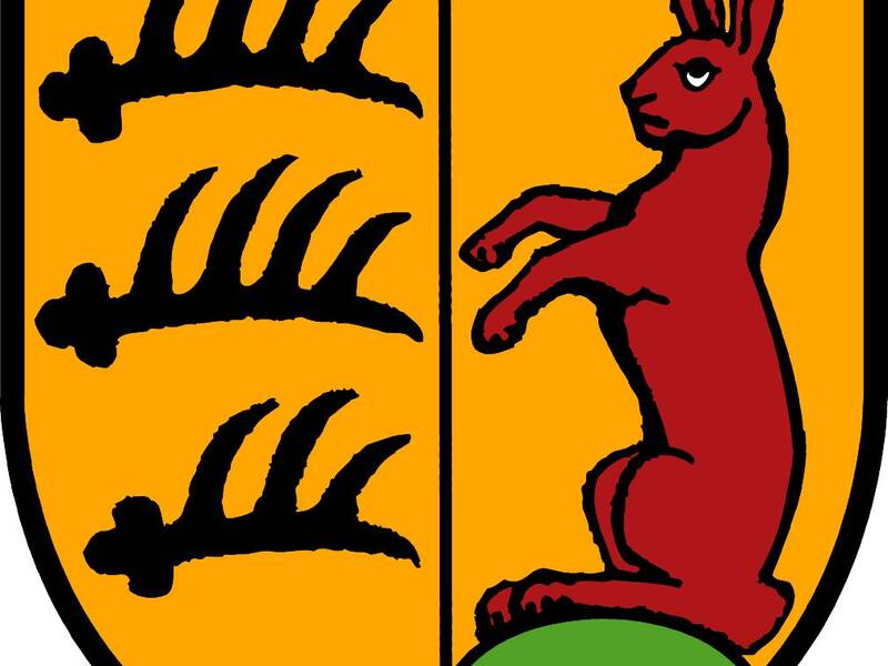 Das Bild zeigt ein Wappen mit drei schwarzen Hirschstangen auf gelbem Grund links und einem aufrechten roten Hasen auf grünem Hügel rechts.