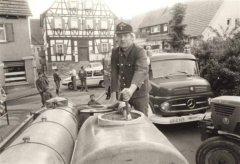 Ein Feuerwehrmann, gekleidet in Uniform, befüllt im Freien einen Tankwagen mit Wasser, während im Hintergrund ein Traktor, ein Feuerwehrauto und ein Fachwerkhaus zu sehen sind.
