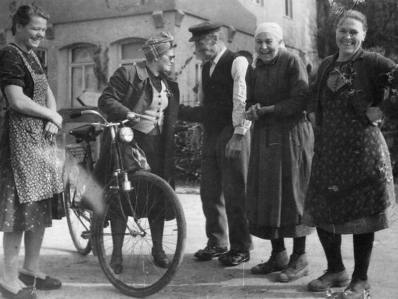 Eine Gruppe von fünf älteren Personen, vier Frauen und ein Mann, steht lachend in einem ländlichen Umfeld neben einem Fahrrad.