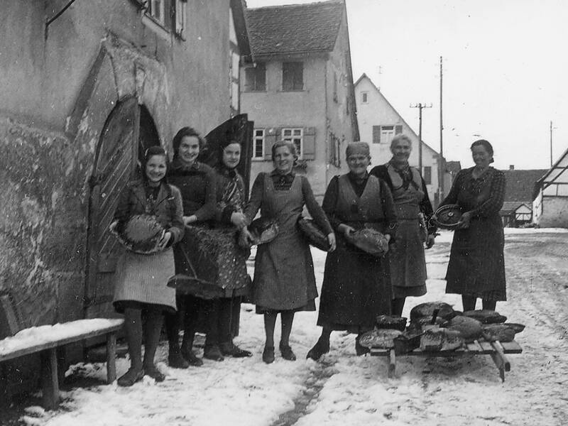 Eine Gruppe von sieben Frauen steht auf einer verschneiten Straße vor einem Gebäude und hält Brote, während weitere Brote auf einem hölzernen Schlitten vor ihnen liegen.