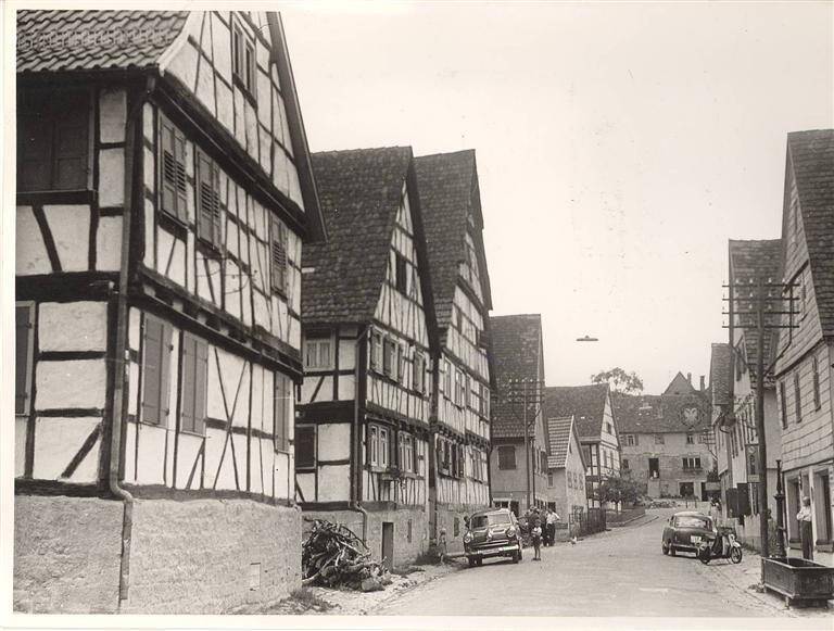 Eine historische Schwarz-Weiß-Fotografie zeigt eine Straße in einem Dorf mit Fachwerkhäusern und zwei geparkten Autos.