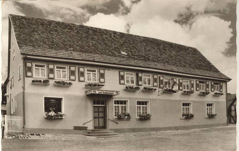 Das Bild zeigt ein zweistöckiges, traditionelles Gasthaus mit Blumenkästen an den Fenstern und einem hölzernen Schild, unter einem bewölkten Himmel.