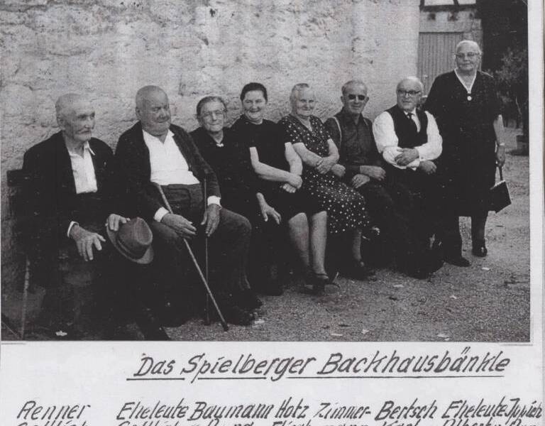 Acht ältere Personen sitzen auf einer Bank vor einer Steinmauer und sind lächelnd der Kamera zugewandt, mit einigen handgeschriebenen Namen am unteren Rand des Bildes.