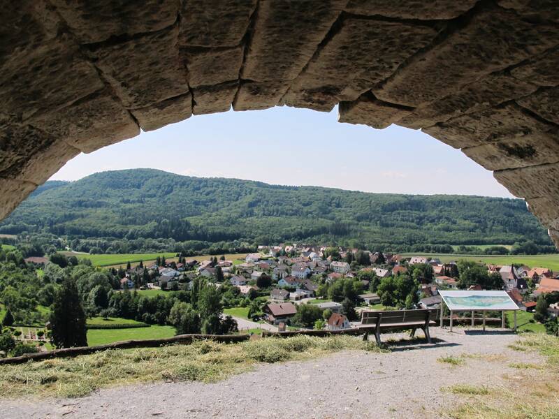 Der Blick aus einer steinernen Höhle zeigt eine grüne Landschaft mit Hügeln und ein Dorf im Tal, ergänzt durch eine Bank und eine Informationstafel im Vordergrund.