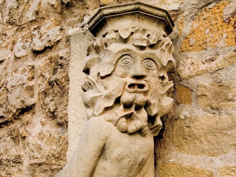 Die steinerne Wasserspeierfigur ist an einer sandfarbenen Mauer befestigt und zeigt einen Mann mit wildem, blättrigem Haar, der in die Hocke geht.