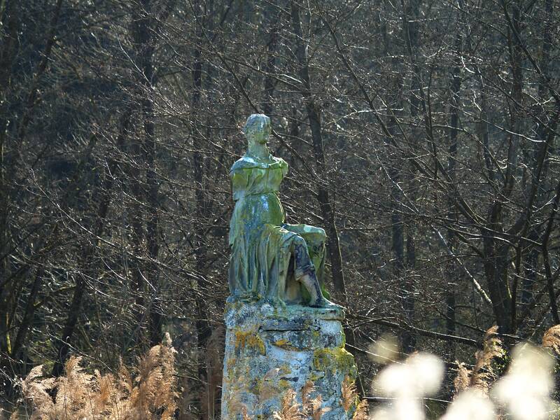 Eine alte, verwitterte Statue steht auf einem Sockel inmitten einer dicht bewachsenen, winterlichen Waldszene, umgeben von hohen, trockenen Gräsern.