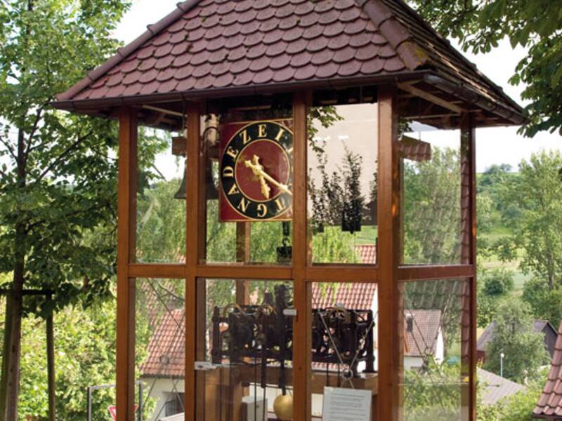 Ein gläserner Pavillon mit einem Ziegeldach beherbergt eine mechanische Uhr und steht neben einem gepflasterten Weg unter Bäumen.