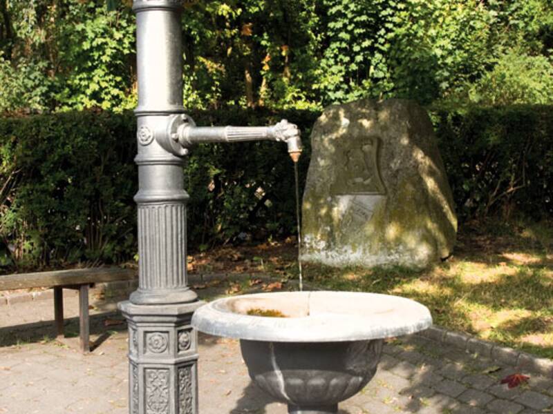 Ein traditioneller Wasserbrunnen aus Metall steht in einem gepflasterten Parkbereich, umgeben von grünen Büschen und einem großen Felsen im Hintergrund.