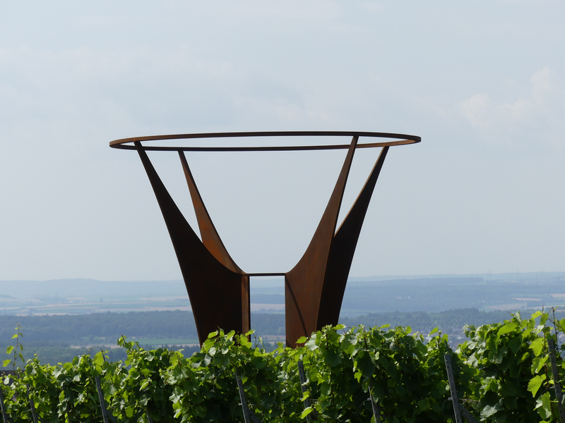 Eine moderne Skulptur in Form eines Kelches aus rostigem Metall steht über einem Weinberg vor einem weiten Landschaftshorizont.