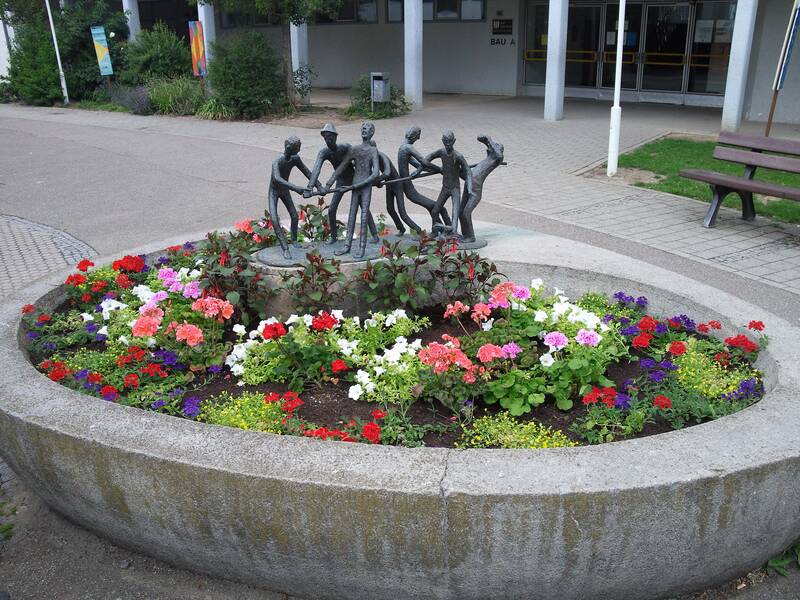 Eine runde Blumenrabatte mit bunten Blumen umgibt eine Bronzeskulptur von Menschen, die sich an den Händen halten.