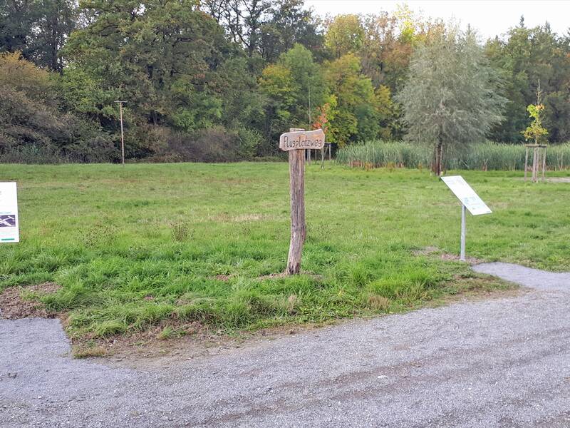 Ein Weg mit mehreren Informationstafeln führt durch eine naturnahe Landschaft mit Bäumen und Sträuchern.