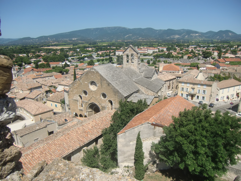 Eine historische Kirche mit mehreren Rundfenstern und einem Glockenturm steht im Zentrum einer mediterranen Stadtlandschaft vor einer Bergkulisse.