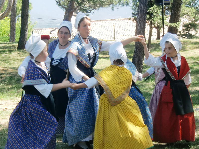 Eine Gruppe von Personen in traditionellen Trachten tanzt im Freien auf einer Wiese unter Bäumen.