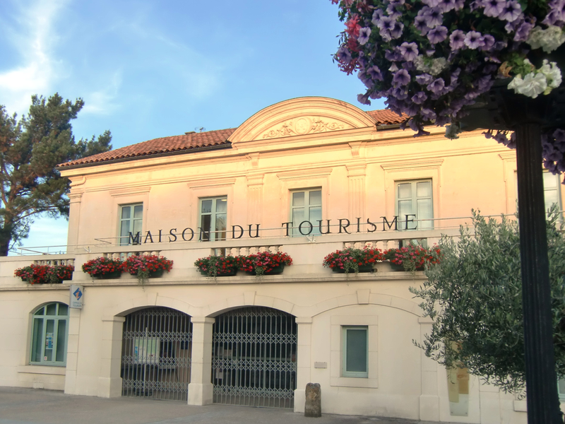 Ein zweistöckiges Gebäude mit der Aufschrift "Maison du Tourisme" und roten Blumenkästen an den Fenstern steht unter blauem Himmel.