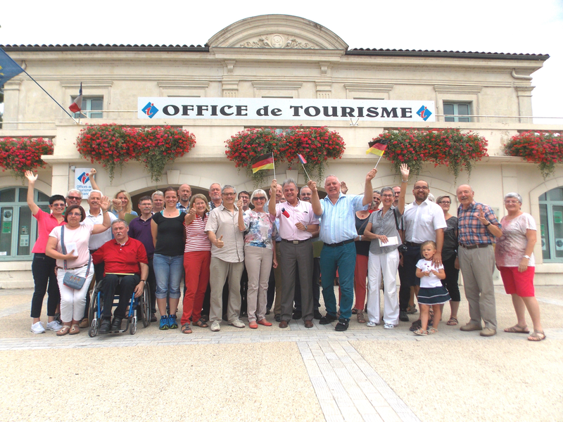 Eine Gruppe von fröhlichen Menschen steht vor einem Gebäude mit der Aufschrift "Office de Tourisme", einige halten Fahnen und winken.