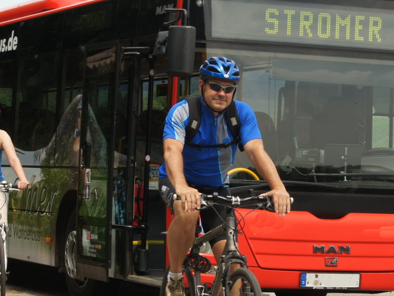 Ein roter Linienbus mit der Aufschrift "Stromer" fährt auf einer Straße in ländlicher Umgebung, während mehrere radfahrende Personen auf einem Radweg daneben unterwegs sind.