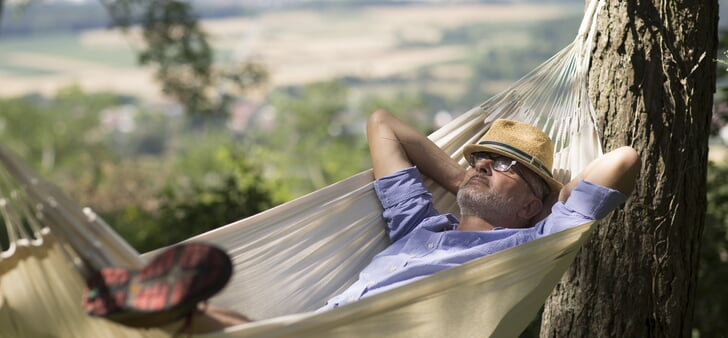 Eine Person mit Hut liegt entspannt in einer Hängematte zwischen Bäumen, mit einer malerischen Landschaft im Hintergrund.