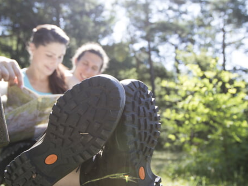 Zwei Personen sitzen auf einer Holzbank im Freien, mit Fokus auf ihre Wanderschuhe im Vordergrund vor einer sonnigen, grünen Naturkulisse.