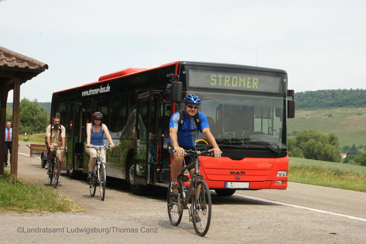 Ein roter Linienbus mit der Aufschrift "Stromer" fährt auf einer Straße in ländlicher Umgebung, während mehrere radfahrende Personen auf einem Radweg daneben unterwegs sind.