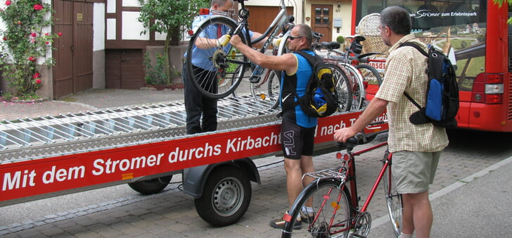 Mehrere Personen laden Fahrräder auf einen ausziehbaren Anhänger mit der Aufschrift "Mit dem Stromer durchs Kirbach- und Weißbachtal", der an einen Bus gekoppelt ist.
