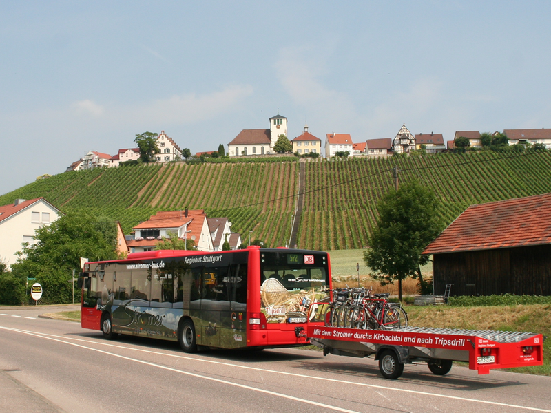 Ein roter Linienbus mit einem Anhänger für Fahrräder fährt eine Landstraße entlang, im Hintergrund sind Weinberge und ein Dorf auf einem Hügel zu sehen.
