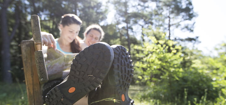 Zwei Personen sitzen auf einer Holzbank im Freien, mit Fokus auf ihre Wanderschuhe im Vordergrund vor einer sonnigen, grünen Naturkulisse.