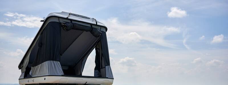 Ein Auto mit einem geöffneten Dachzelt steht vor einer weiten, grünen Landschaft unter einem teilweise bewölkten Himmel.