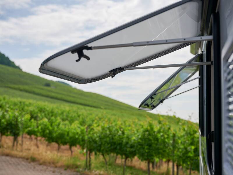 Die Nahaufnahme zeigt ein Wohnmobilfenster, das geöffnet ist, mit einer Landschaft aus Weinbergen im Hintergrund.