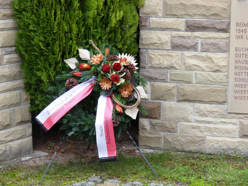 Ein Kranz aus bunten Blumen mit zwei Schleifen steht vor einer Steinmauer mit einer Gedenktafel.