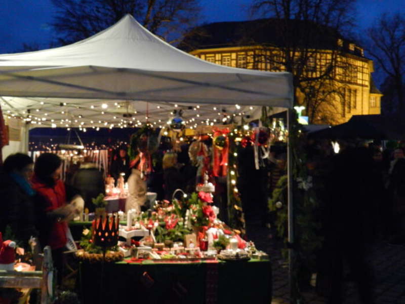 Ein gut beleuchteter Weihnachtsmarktstand mit vielen dekorativen Artikeln und zwei Personen steht vor einem großen, erleuchteten Gebäude in der Abenddämmerung.