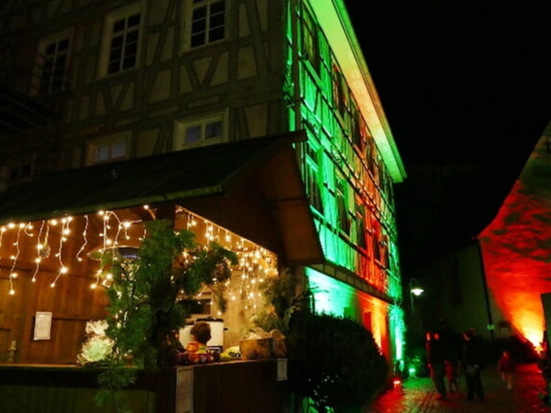 Ein Fachwerkhaus und ein Marktstand sind nachts festlich mit Lichterketten und grün-roter Beleuchtung geschmückt.