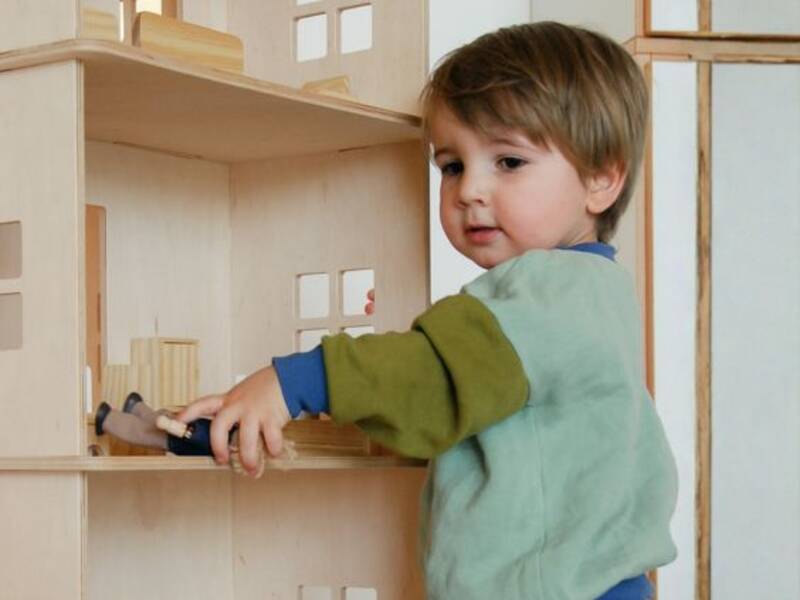 Ein kleines Kind spielt mit einem Holzspielzeughaus und hält dabei einen Hammer.