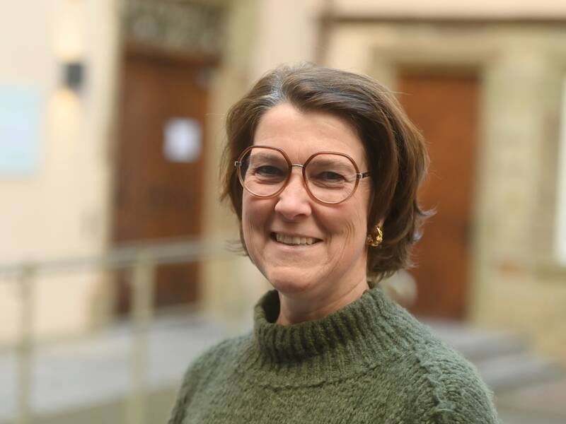 Frau Toussaint-Prella mit Brille und grünem Pullover lächelt in die Kamera, im Hintergrund sind unscharfe Gebäude erkennbar.