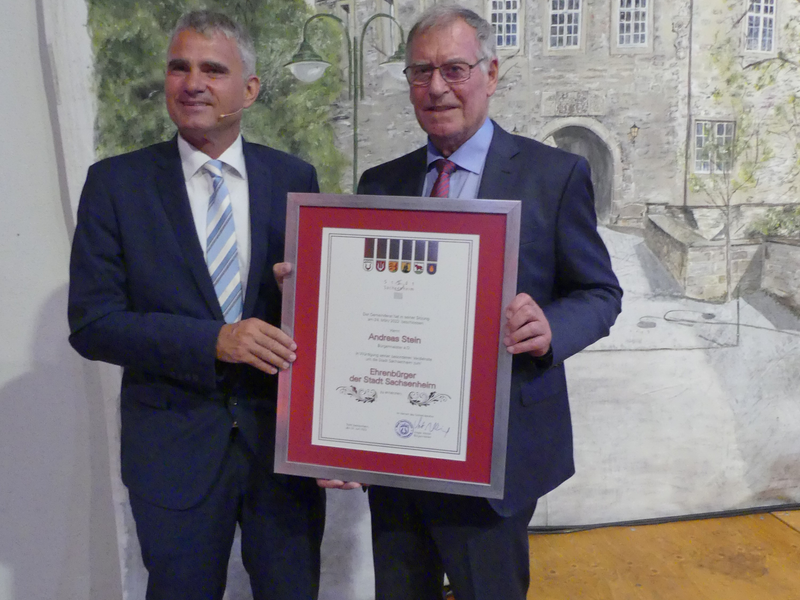 Bürgermeister Albrich und Herr Stein a.D. halten eine gerahmte Urkunde vor einer Wand mit einem Gemälde eines Fachwerkhauses.
