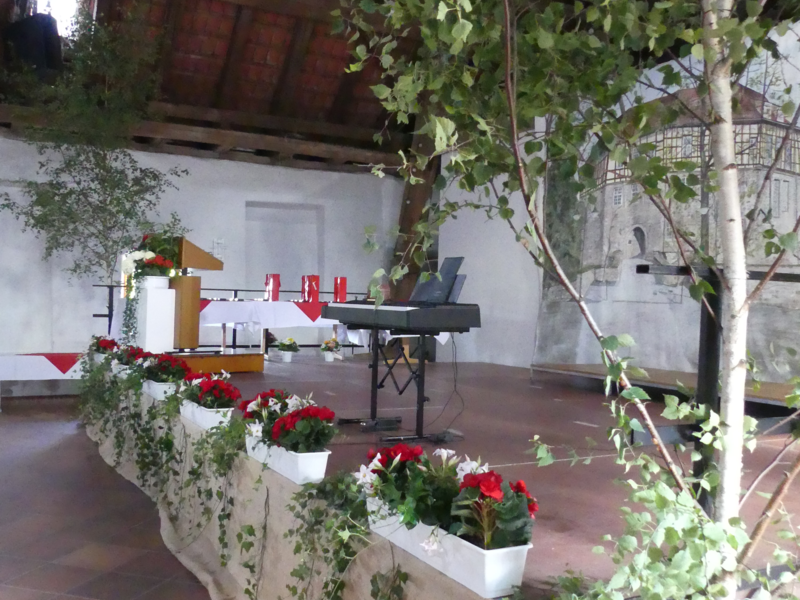 Die Indoor-Szene zeigt einen mit Pflanzen und Blumen geschmückten Bereich, darunter ein Keyboard und ein Pult, umgeben von Backsteinwänden und Holzbalken unter einem Dach.