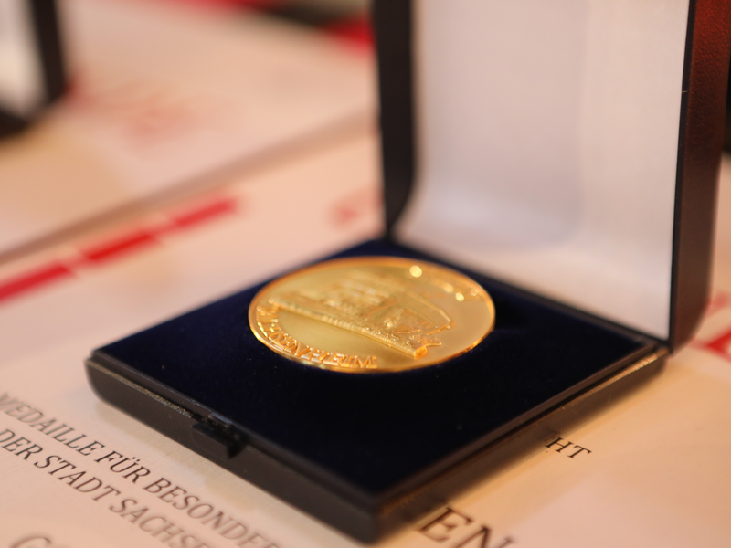 Eine goldfarbene Medaille liegt in einer schwarzen Box mit blauer Innenauskleidung, die auf einem Dokument platziert ist.