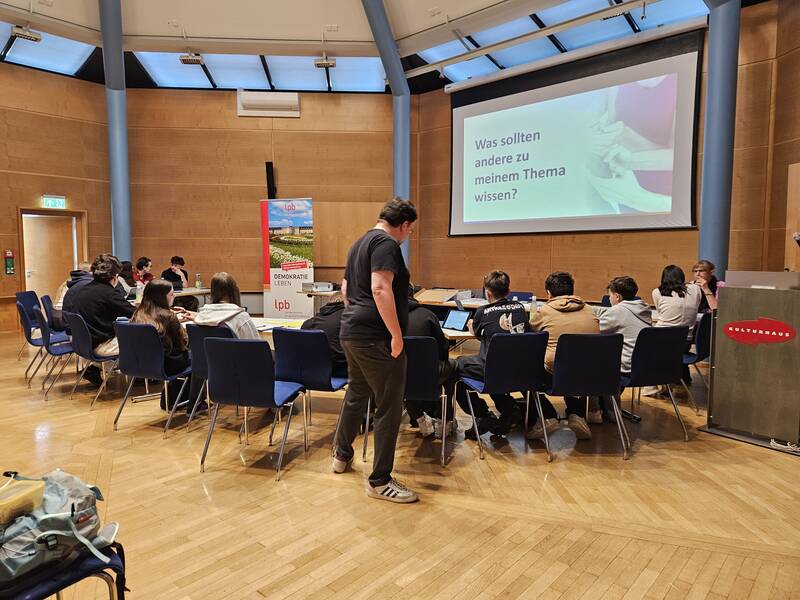 Ein Dutzend Menschen sitzt in einem runden Konferenzraum und schaut auf eine Leinwand mit der Frage "Was sollten andere zu meinem Thema wissen?".