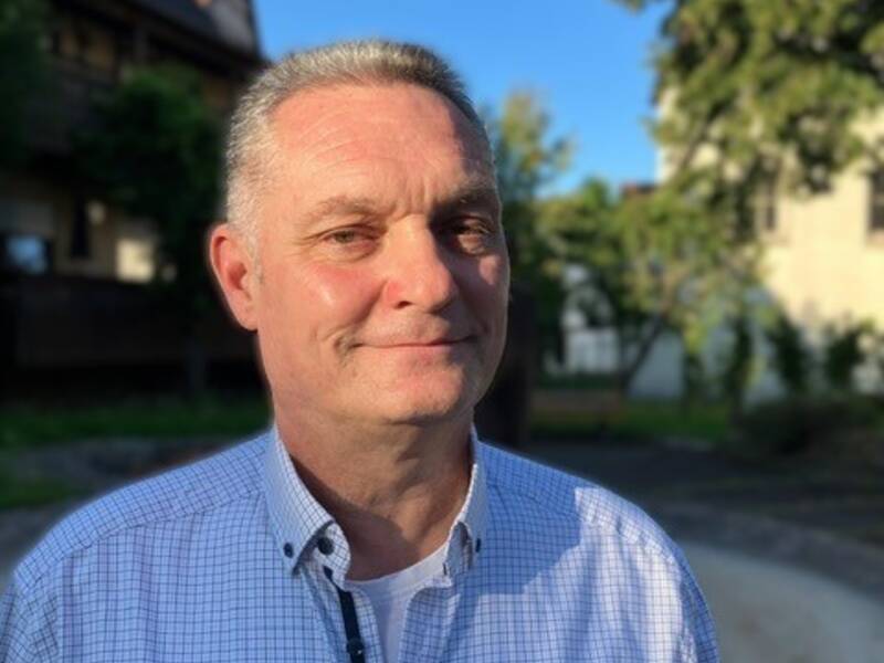 Herr Weiß im karierten Hemd steht draußen in einem sonnigen Garten vor einem Haus.