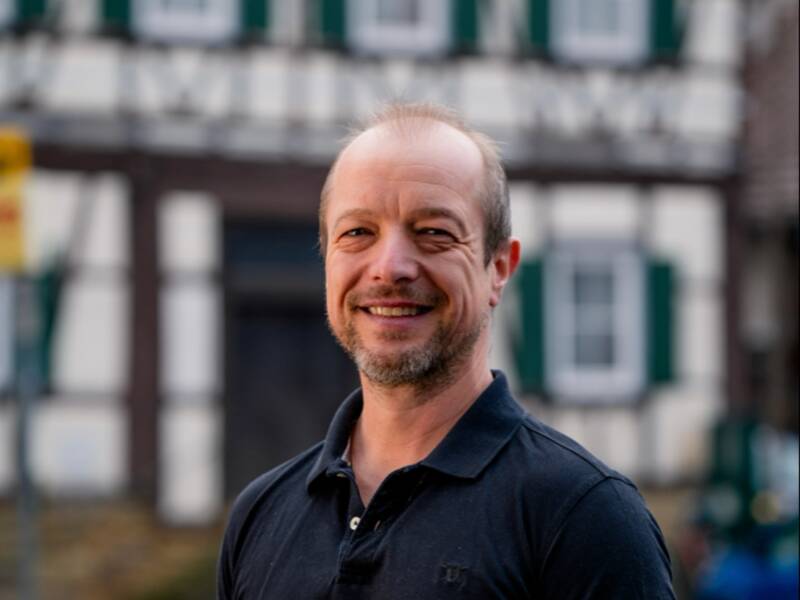Herr André Lux steht in einem schwarzen Poloshirt vor einem Fachwerkhaus.