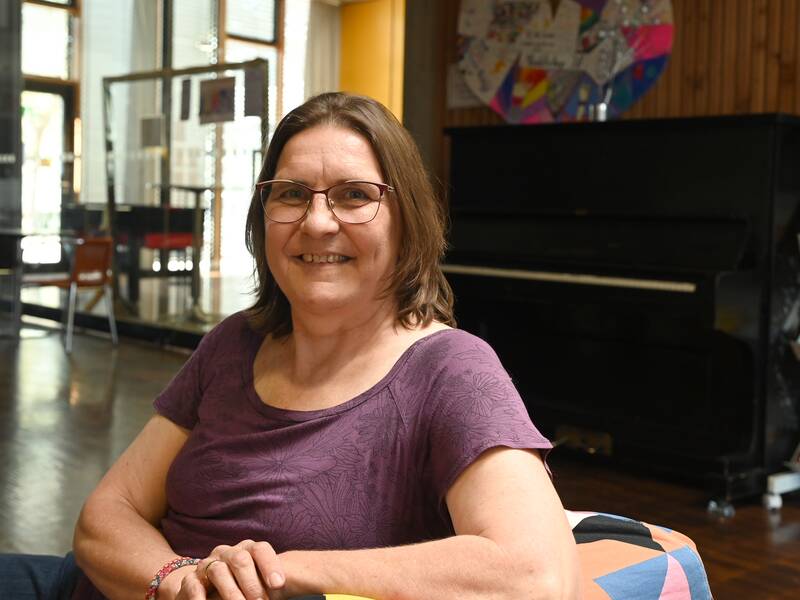 Frau Lange mit Brille und lila Oberteil sitzt in einem Raum mit Holzboden und einem Klavier im Hintergrund.