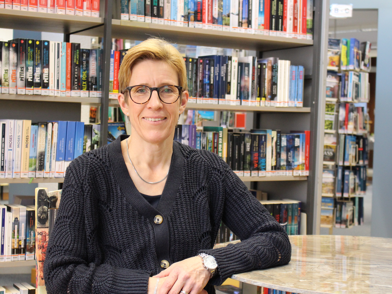 Eine Person mit Brille und schwarzem Strickpullover sitzt an einem Tisch in einer Bibliothek vor einem Regal voller Bücher.