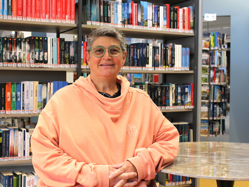Eine Person mit Brille und rosa Kapuzenpullover sitzt in einer Bibliothek vor einem Tisch, umgeben von vollen Bücherregalen.