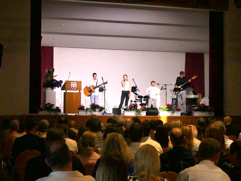 Auf einer Bühne spielt eine Akustikband der Musikschule aus fünf Musikern vor einem sitzenden Publikum.