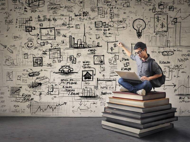 Ein Junge der auf einem Stapel Bücher sitzt arbeitet am Laptop und zeigt auf die Wand hinter sich die mit verscheidenen Symbolen und Grafiken bemalt ist