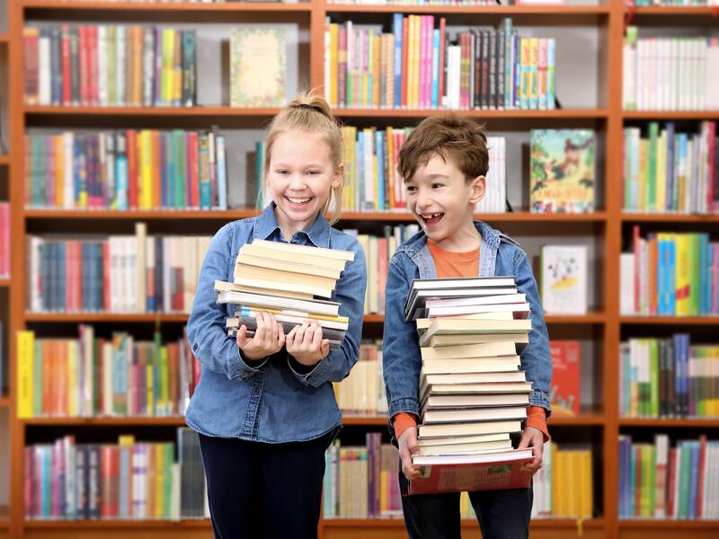 Zwei Kinder halten je einen Stapel Bücher und lachen, sie stehen vor einem bunten Bücherregal